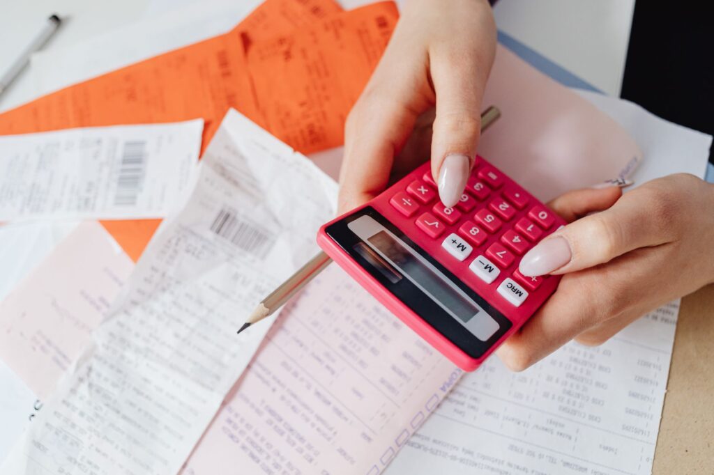 hands using a pink calculator to manage expenses amidst various receipts and documents.