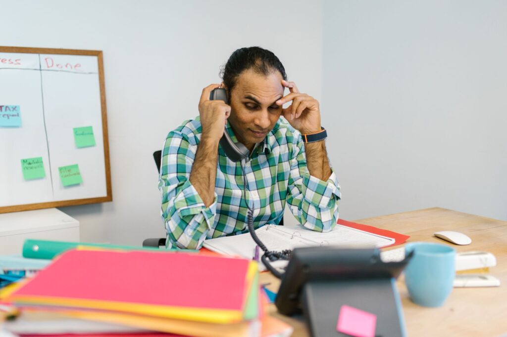 a businessman feeling stressed while talking on the phone, surrounded by paperwork in a busy office setting.