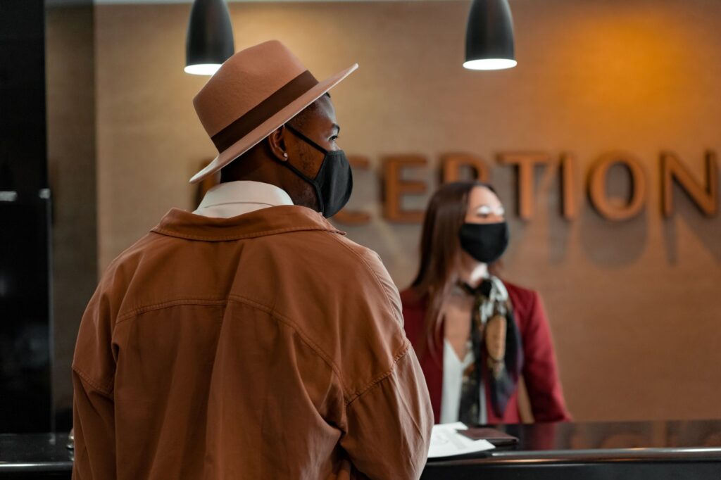 man in mask at hotel reception during pandemic, interacting with masked receptionist.