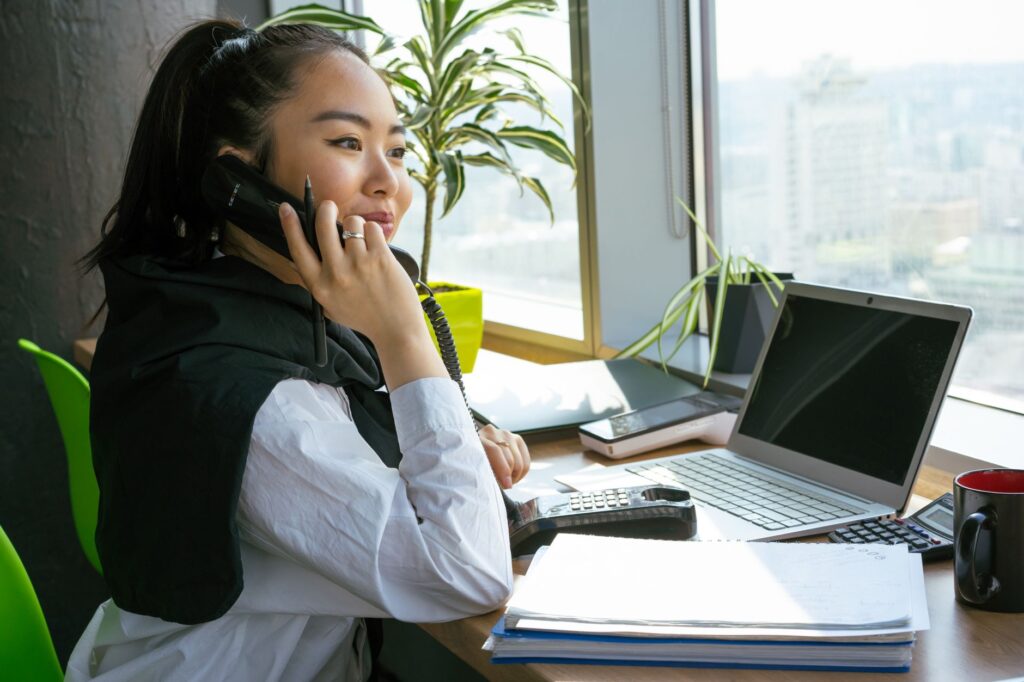 woman on a phone call surrounded by a laptop, papers, and a potted plant in a bright office setting.